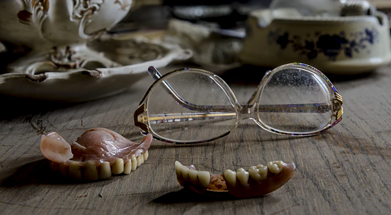 A pair of dentures on a wooden table next to reading glasses
